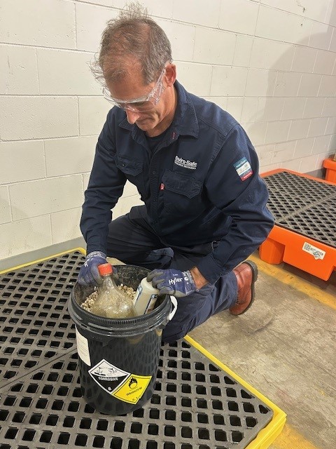Enviro-Safe technician wearing safety glasses and gloves handling containers in a hazardous waste pail on a spill containment platform.