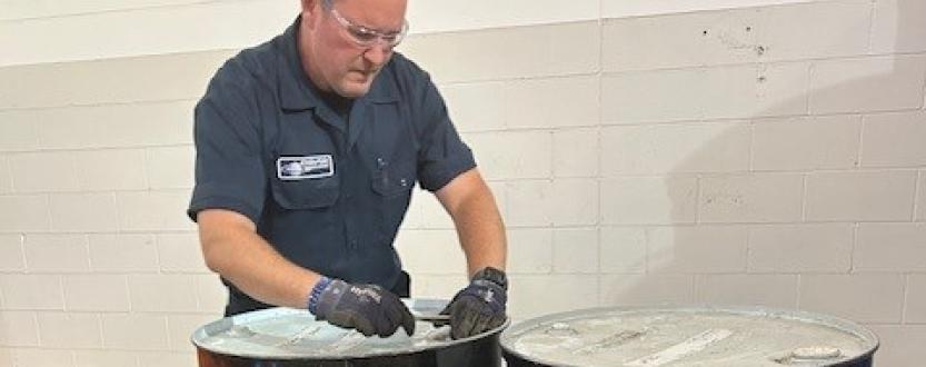 Gloved Enviro-Safe technician working on the lid of a black drum beside a second drum.